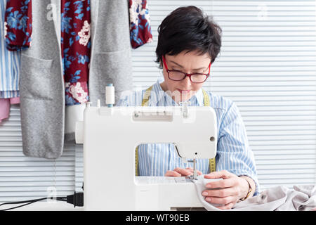 Moda sarta in occhiali lavora al negozio di sartoria con impianto elettrico macchina per cucire industriale Foto Stock