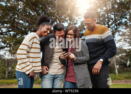 Gruppo di giovani multi razziale amici in piedi insieme guardando al telefono mobile nel parco sorridendo - caldo all'aperto Foto Stock