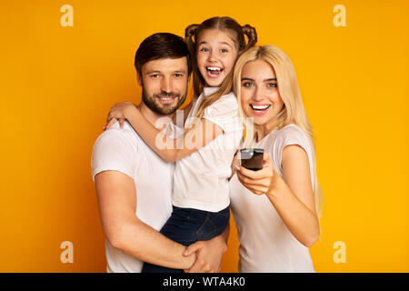 Famiglia guarda la TV canali di commutazione per divertirsi insieme, sfondo giallo Foto Stock