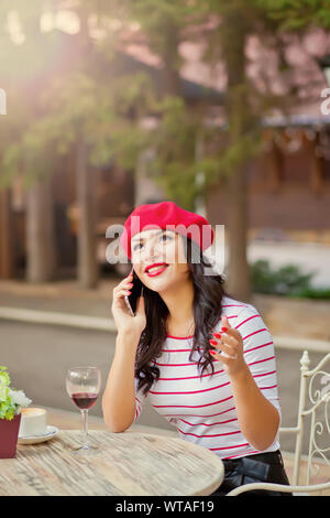 Bella bruna donna in un cappuccio rosso e striato t-shirt sorridente, bere vino rosso in outdoor cafe e parlando al telefono Foto Stock