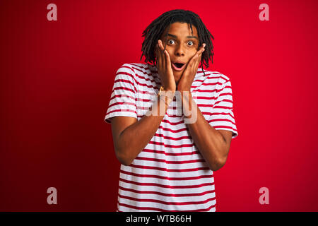 Giovani afro uomo con dreadlocks a strisce che indossa la t-shirt in piedi isolato su sfondo rosso paura e scioccato, sorpresa e stupita espressione con h Foto Stock