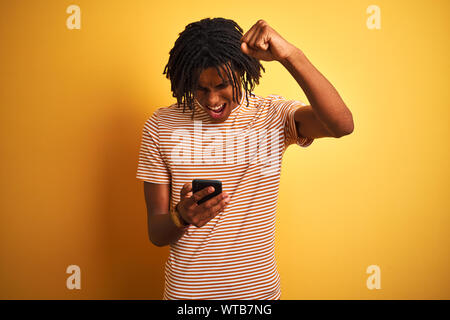 Afro-americano con dreadlocks utilizzando lo smartphone isolato su sfondo giallo infastiditi e frustrato grida con rabbia, crazy e urla con Foto Stock