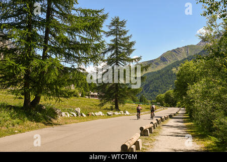 Vista panoramica di un paesaggio di montagna con un paio di ciclisti su una strada stretta tra prati e foreste in estate, Val Ferret, Courmayeur, Alpi Italia Foto Stock