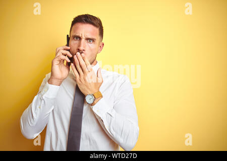 Bel giovane uomo d affari parlando al telefono giallo su sfondo isolato coprono la bocca con la mano sconvolto con vergogna per errore, espressione di f Foto Stock