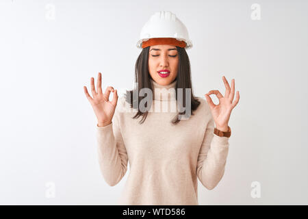 Giovane architetto cinese donna che indossa il casco di protezione isolati su sfondo bianco relax e sorridente con gli occhi chiusi facendo gesto di meditazione con Foto Stock
