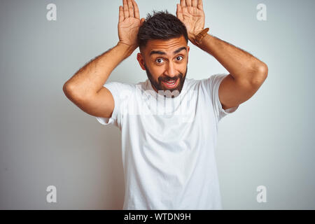 Giovani indiani uomo che indossa la t-shirt in piedi isolato su sfondo bianco facendo orecchie di coniglietto gesti con le mani le palme cercando cinico e scettico. Oriente Foto Stock