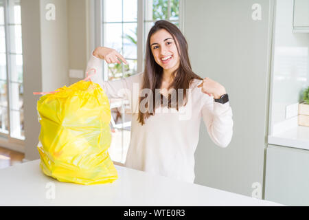 Bella giovane donna portare fuori l'immondizia da il contenitore di rifiuti guardando fiduciosi con il sorriso sul volto, rivolto a se stessi con le dita in fieri e Foto Stock