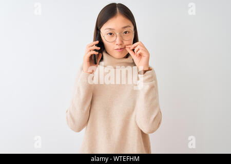 Giovane donna cinese parlando sullo smartphone isolato su sfondo bianco bocca e le labbra chiuse come zip con le dita. Segreto e silenzioso, tabù parlando Foto Stock