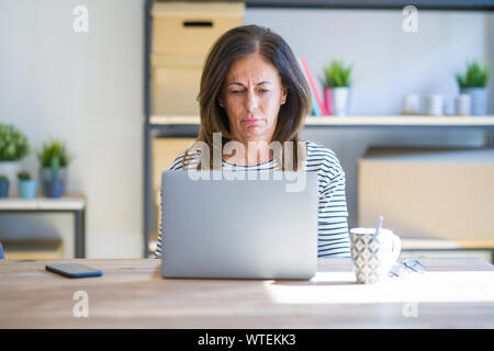 La mezza età senior donna seduta a tavola in casa a lavorare utilizzando il computer portatile premuto e la preoccupazione per la sofferenza, gridando arrabbiato e paura. Triste expr Foto Stock