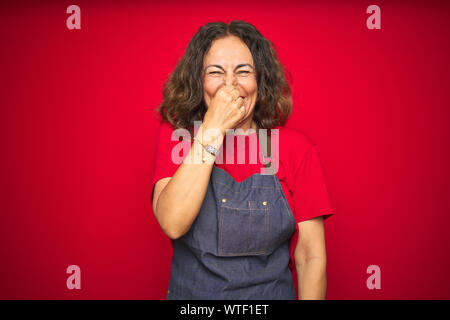 La mezza età senior donna che indossa il grembiule uniforme su rosso sfondo isolato annusare qualcosa di maleodorante e disgustoso, intollerabile odore, apnea Foto Stock