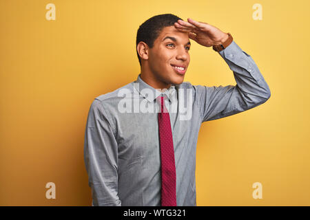 Bel giovane imprenditore arabo indossando camicia e cravatta isolato su sfondo giallo molto felice e sorridente guardando lontano con la mano sopra la testa. Il mare Foto Stock
