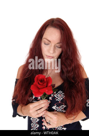 Un triste guardando la donna con lunghi capelli rossi tenendo una rosa rossa in mano guardando giù, stando in piedi in un abito nero isolato per sfondo bianco Foto Stock