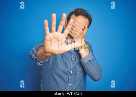 Bella la mezza età senior uomo con i capelli grigi isolati su sfondo blu che copre gli occhi con le mani e facendo arrestare gesto con il triste e paura expressi Foto Stock