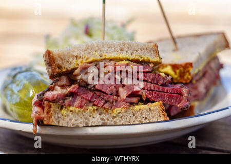 Sandwich Pastrami tagliate a metà sul pane di segale con senape su una piastra bianca su una tavola di legno come visto da sopra. Coleslaw e aneto sottaceto sul lato. Foto Stock