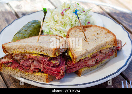 Sandwich Pastrami tagliate a metà sul pane di segale con senape su una piastra bianca su una tavola di legno come visto da sopra. Coleslaw e aneto sottaceto sul lato. Foto Stock