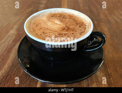 Chai Latte in una tazza nera sul piattino servito su legno scuro ha sottolineato cafe tabella. Popolare bevanda di tè. Vista laterale. Foto Stock