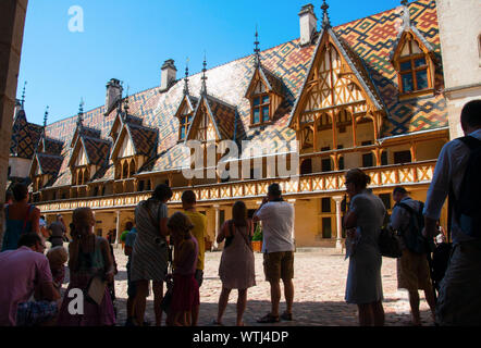 Visite turistiche turisti in piedi nell'ombra presso il cortile dell'Hospices de Beaune con la sua splendidamente decorate tetto. Beaune, Borgogna, Francia. Foto Stock