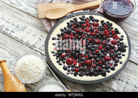 Torta di bacche con ciliegia, ribes, mora, mirtillo con le stoviglie e gli ingredienti di colore bianco su un tavolo di legno. Cheesecake. Foto Stock