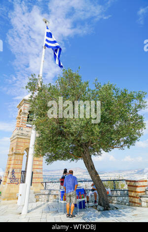 Atene GRECIA - luglio14 2019; bevande fredde venditore sotto l'ulivo e pennone dal campanile alla sommità del Monte Lycabettus Foto Stock