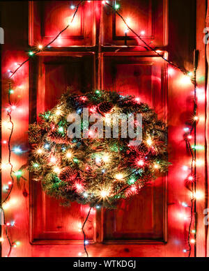 Una ghirlanda di Natale i rami degli alberi con i coni appeso sulla porta anteriore. Notte decorazione della porta con una luminosa ghirlanda luminosa, una corona di abeti Foto Stock