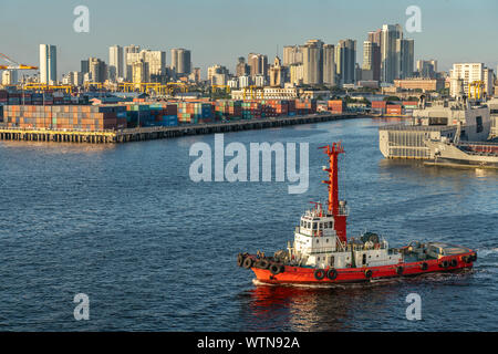 Manila, Filippine - Marzo 5, 2019: sud Porto crepuscolo serale. Red rimorchiatore fino anteriore. Le navi della marina militare, un molo pieno di contenitori di spedizione nel retro. L Foto Stock