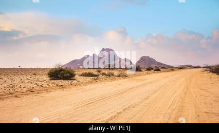 Un vasto paesaggio africano, con una strada sterrata che corre attraverso aride pianure del deserto del Namib verso le vette di granito di Spitzkoppe, Namibia. Foto Stock