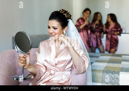 Sposa mattina. La sposa tiene un piccolo specchio nelle sue mani e guarda a lei amiche dietro Foto Stock