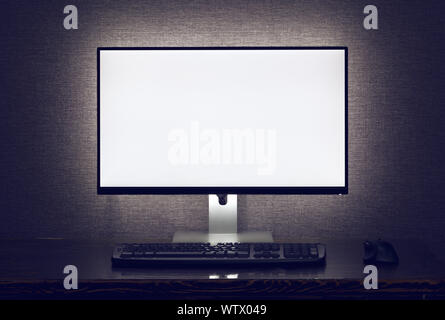 Monitor in bianco con tastiera e mouse con retroilluminazione oltre il muro grigio in dark room Foto Stock