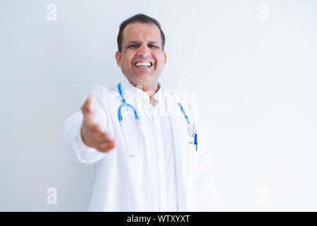 La mezza età medico uomo che indossa lo stetoscopio e il rivestimento di medici su sfondo bianco sorridente cordiale offrendo stretta di mano come saluto e accogliente. Successo Foto Stock