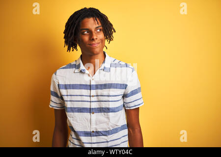 Afro-americano con dreadlocks indossando camicia a righe isolate su sfondo giallo sorridente guardando al lato e lo sguardo lontano pensando. Foto Stock
