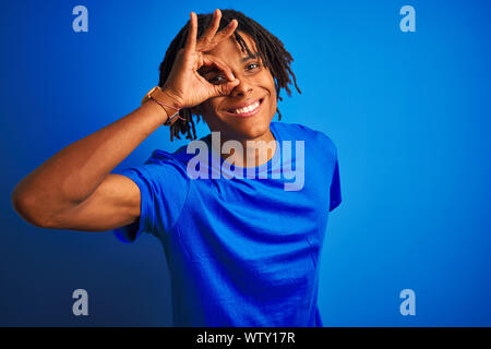 Afro-americano con dreadlocks che indossa la t-shirt in piedi isolato su sfondo blu facendo ok gesto con la mano sorridendo, occhio guardando attraverso finge Foto Stock