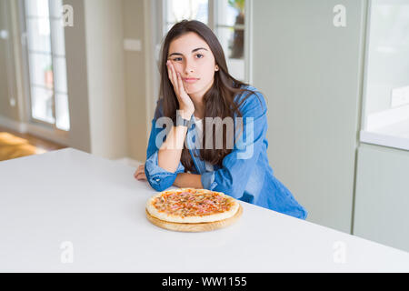 Bella giovane donna di mangiare in casa una gustosa pizza al pensiero di cucina cercando stanco ed annoiato con depressione problemi con bracci incrociati. Foto Stock