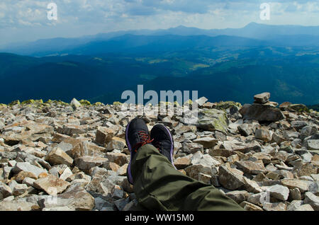 L uomo per le gambe nella tracciatura di sneakers in cima alla montagna contro lo sfondo di catene montuose e cielo blu Foto Stock