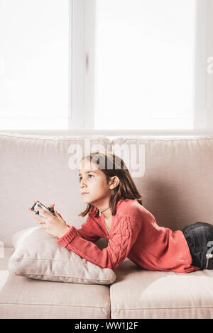 Niña jugando en casa con la videoconsola tumbada en el divani Foto Stock