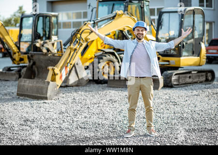 Ritratto di un bel builder in piedi sul terreno aperto del negozio con macchinari pesanti per la costruzione Foto Stock