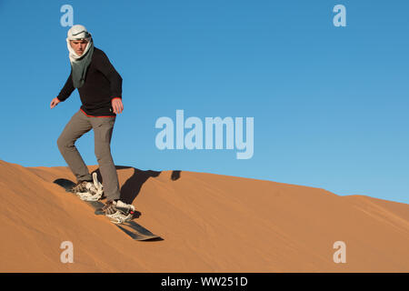 Un uomo con un velo praticano sandboarding nelle dune del deserto di Erg Chebbi Foto Stock