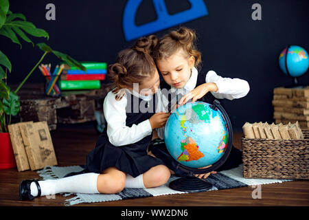 Poco twin ragazze in uniforme scolastica con lente di ingrandimento per esaminare il mondo stando seduti sul pavimento durante la lezione di geografia Foto Stock