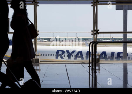 Passeggeri a piedi con i bagagli nel terminal dell'aeroporto, con Ryanair in aereo in background Foto Stock