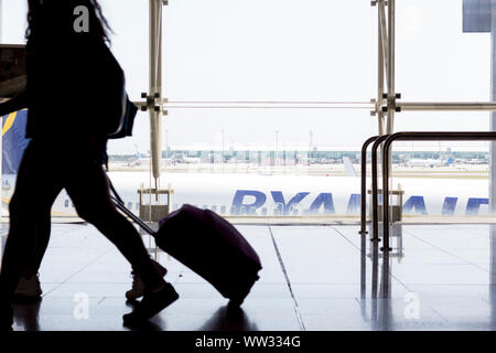 Passeggeri a piedi con i bagagli nel terminal dell'aeroporto, con Ryanair in aereo in background Foto Stock