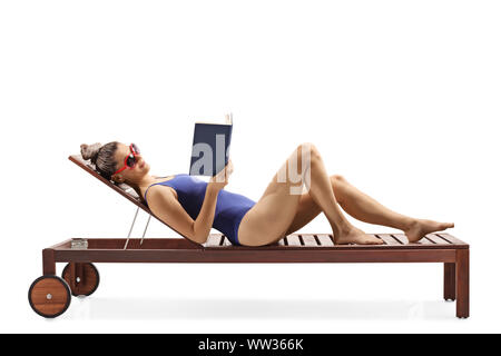 Lunghezza piena ripresa di profilo di una giovane donna in un costume da bagno che giace su una sedia a sdraio e readng un libro isolato su sfondo bianco Foto Stock