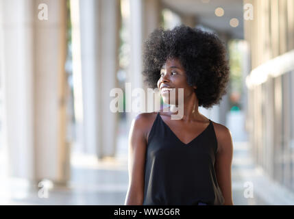 Sorridente giovane donna nera a piedi nella città guardando al lato, la bella giornata di sole Foto Stock