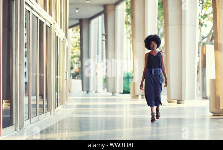 Un giovane alla moda donna Afroamericana con fiducia a camminare per il corridoio al di fuori dell'edificio finanziario Foto Stock