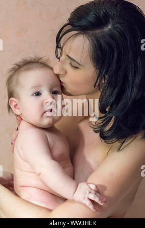 Giovane madre bella bruna senza vestiti tenendo le braccia e baciare il suo piccolo figlio Foto Stock