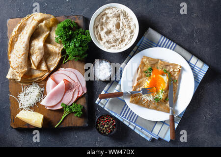 Crêpe bretone, grano saraceno Galette Bretonnes con lato soleggiato in uovo, il formaggio, il prosciutto su una piastra bianca su una tabella di calcestruzzo con ingredienti su una boa di taglio Foto Stock