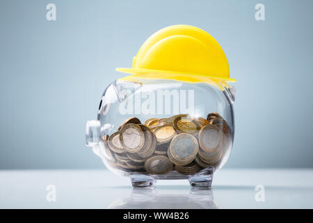 Close-up di Casco giallo su trasparente di salvadanaio sulla scrivania bianca contro lo sfondo blu Foto Stock