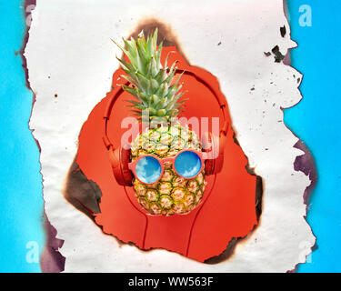 Funny ananas blu dipinta in rosso in occhiali da sole e auricolari. Appartamento laici corallo su carta a colori con cerchi. Foto Stock