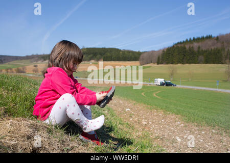 5-anno-vecchia ragazza con giacca rossa seduta al fieldedge, mettendo sulle scarpe Foto Stock