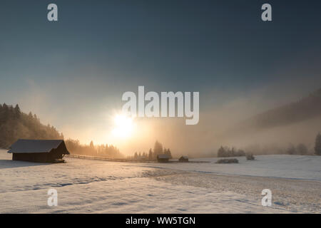 Capanna in coperta di neve inverno alpino paesaggio vicino a Garmisch Partenkirchen. Sullo sfondo il Rising Sun nella nebbia mattutina con altre capanne di legno. Foto Stock