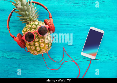 Funny ananas in occhiali da sole e auricolari dipinto di blu. Lay piatto su carta rossa sfondo con sfere di soffici e retrò nastri audio. Foto Stock