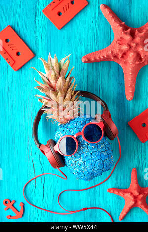 Tanga carattere di ananas in rosso gli occhiali da sole e auricolari ascolta la musica. Testurizzato rustico in legno pannelli sfondo dipinto di blu, vista dall'alto, fl Foto Stock